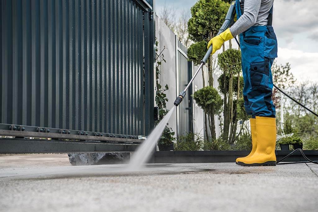 Professional in yellow boots and blue overalls using a pressure washer on a concrete driveway near a metal gate.